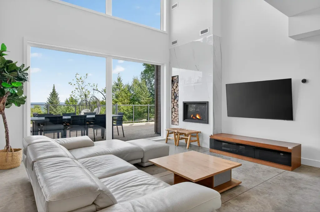 Salon moderne lumineux avec canapé d'angle blanc, cheminée, grande baie vitrée sur terrasse et arbres.